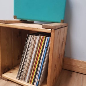May include: A wooden record storage crate with a teal suitcase on top. The crate is filled with vinyl records.