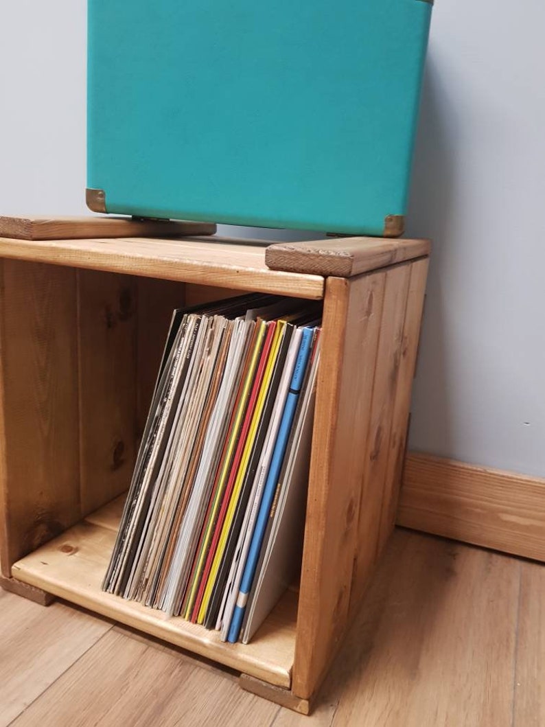 May include: A wooden record storage crate with a teal suitcase on top. The crate is filled with vinyl records.