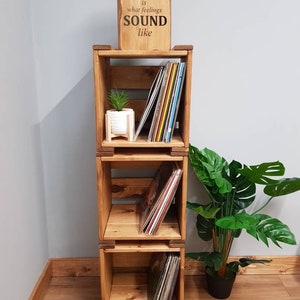 May include: A wooden record shelf with three tiers, each tier holding a stack of vinyl records. The shelf is made of natural wood and has a rustic look. A small potted plant sits on the top tier. A wooden sign above the shelf reads "MUSIC is what feelings SOUND like".