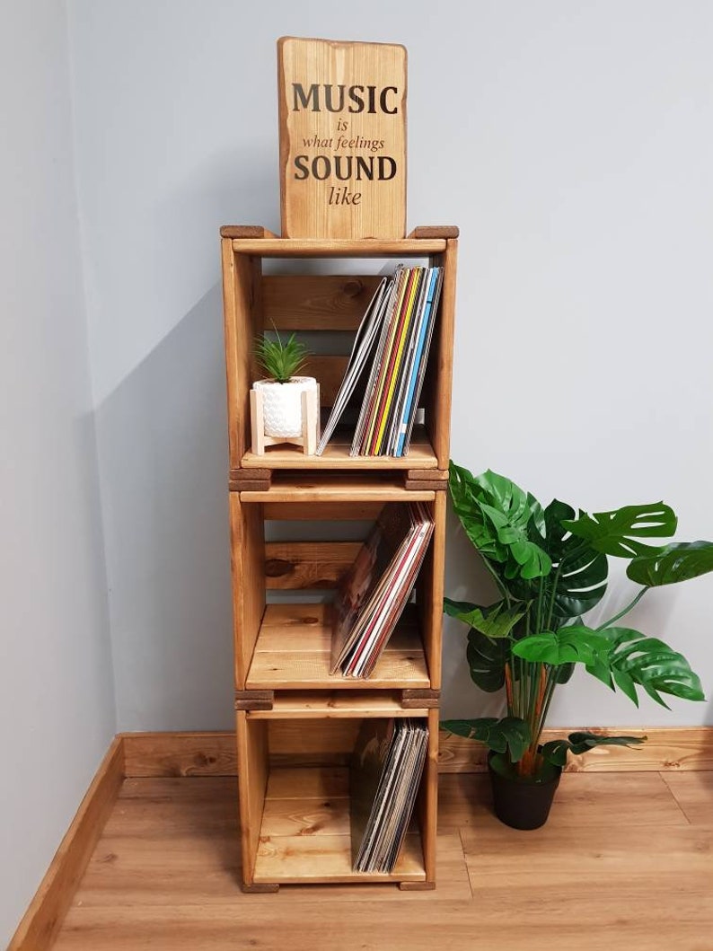May include: A wooden record shelf with three tiers, each tier holding a stack of vinyl records. The shelf is made of natural wood and has a rustic look. A small potted plant sits on the top tier. A wooden sign above the shelf reads "MUSIC is what feelings SOUND like".