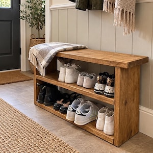 Puede incluir: Un zapatero rectangular de madera con dos estantes, con un acabado de veta de madera marrón medio. El estante contiene varios pares de zapatillas y botas, y una manta a cuadros gris y blanca está colocada sobre la superficie superior.