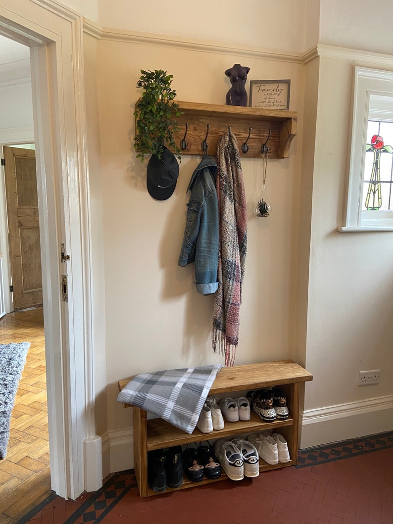 May include: A wooden coat rack with hooks and a shelf, featuring a plant, a hat, a jacket, and a scarf. Below the rack is a wooden shoe rack with two shelves, holding a variety of shoes. The wall is painted a light cream colour. The text on the sign reads "Family is not an important thing. It's everything."