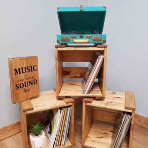 May include: Three wooden crates with shelves, each holding vinyl records. A teal record player sits on top of the crate in the centre. A wooden sign reads "MUSIC what is feelings SOUND like".