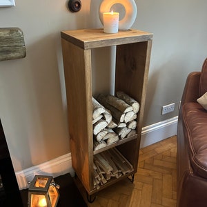 May include: A wooden log storage rack with two shelves. The rack is stained a dark brown color and has black metal hairpin legs. The top shelf is empty and has a white ceramic vase with a candle inside. The bottom shelf is filled with logs.