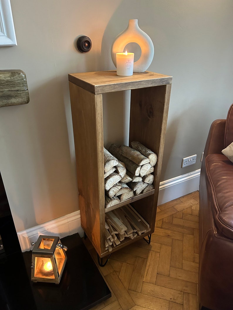 May include: A wooden log storage rack with two shelves. The rack is stained a dark brown color and has black metal hairpin legs. The top shelf is empty and has a white ceramic vase with a candle inside. The bottom shelf is filled with logs.