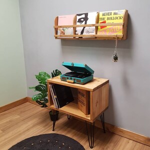 Puede incluir: Un soporte de tocadiscos de madera con un estante para discos de vinilo. El soporte tiene patas de horquilla negras y est&aacute; sentado sobre una alfombra negra y marr&oacute;n. Un tocadiscos turquesa est&aacute; en la parte superior del soporte. Un estante de madera con discos de vinilo est&aacute; montado en la pared detr&aacute;s del soporte.
