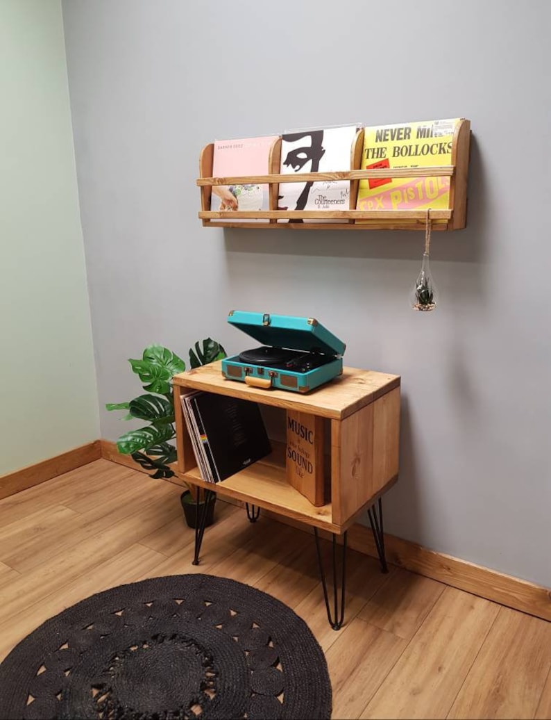 Puede incluir: Un soporte de tocadiscos de madera con un estante para discos de vinilo. El soporte tiene patas de horquilla negras y est&aacute; sentado sobre una alfombra negra y marr&oacute;n. Un tocadiscos turquesa est&aacute; en la parte superior del soporte. Un estante de madera con discos de vinilo est&aacute; montado en la pared detr&aacute;s del soporte.