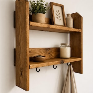 May include: A two-tiered wooden shelf with a warm brown finish. The top shelf holds a potted plant, a framed botanical print, and a small vase. The lower shelf features a bowl, a jar, and two black hooks. A tan towel hangs from the hooks.