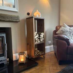 May include: A wooden log storage rack with two shelves, featuring a rustic brown finish and black hairpin legs. The rack is filled with stacked logs and is placed in a living room setting with a fireplace and a leather sofa.