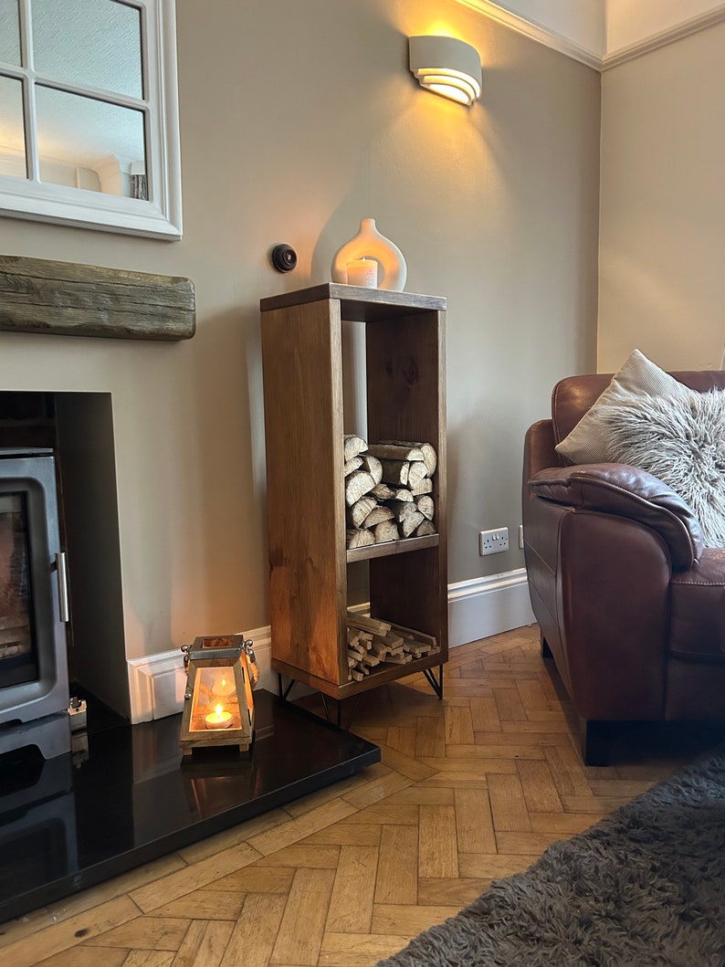 May include: A wooden log storage rack with two shelves, featuring a rustic brown finish and black hairpin legs. The rack is filled with stacked logs and is placed in a living room setting with a fireplace and a leather sofa.