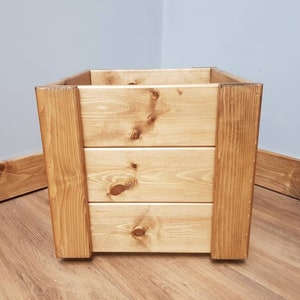 May include: A square wooden planter box with a natural wood finish. The planter has three horizontal slats on the front and sides, creating a rustic look.