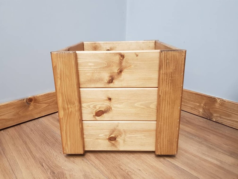 May include: A square wooden planter box with a natural wood finish. The planter has three horizontal slats on the front and sides, creating a rustic look.