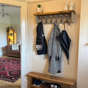 May include: A wooden coat rack with a shelf and hooks, a wooden bench with two shelves, and a wicker basket. The coat rack has a grey painted finish and the bench has a natural wood finish. There are shoes on the shelves of the bench, a coat hanging on a hook, and an umbrella hanging on another hook.
