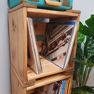 May include: A wooden record storage unit with three shelves. The shelves are stacked on top of each other and are made of natural wood. The unit is filled with vinyl records. The top shelf has a record with a band on the cover. The record is titled "The Beatles 1962-1970".
