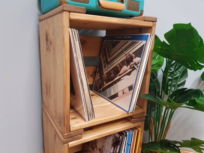 May include: A wooden record storage unit with three shelves. The shelves are stacked on top of each other and are made of natural wood. The unit is filled with vinyl records. The top shelf has a record with a band on the cover. The record is titled "The Beatles 1962-1970".