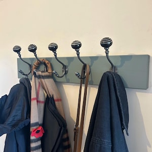 May include: A wooden coat rack with five black hooks and a light green painted finish. A scarf, a jacket, and a leather strap are hanging on the hooks.
