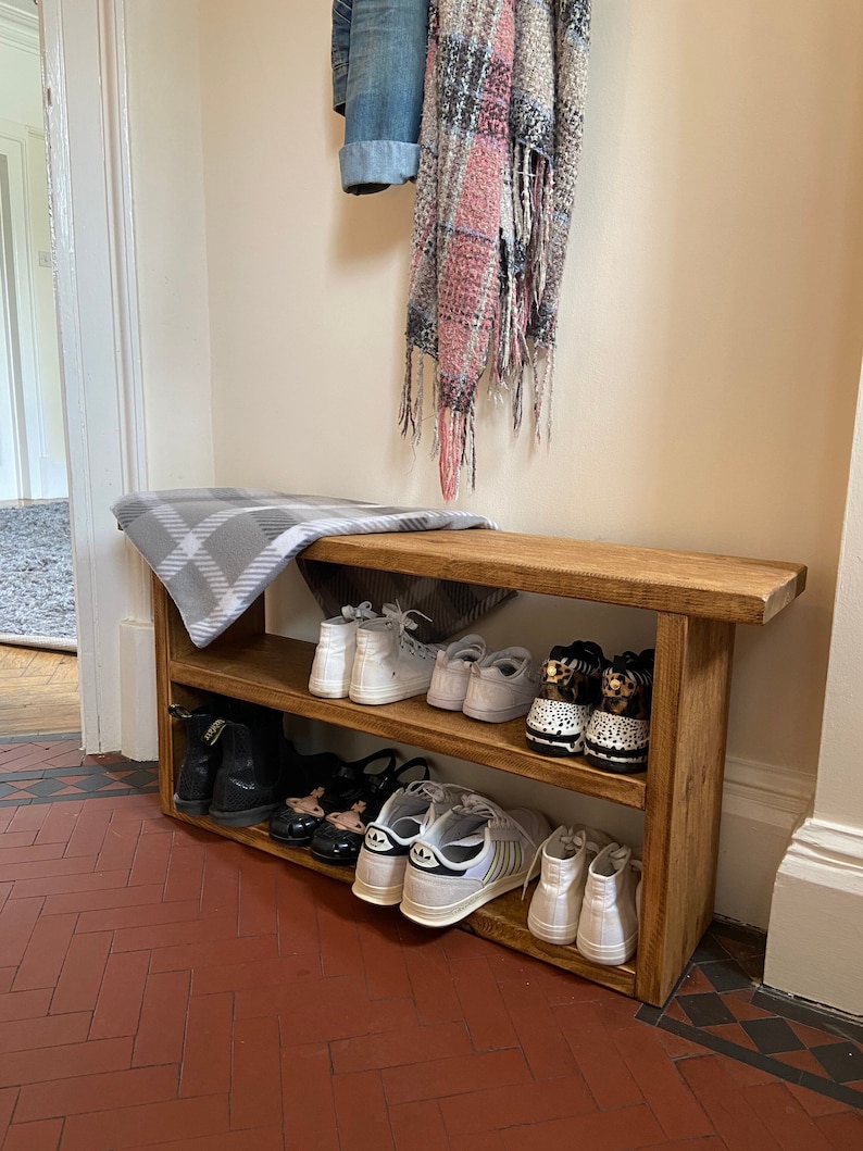 May include: A wooden shoe rack with two shelves, holding a variety of footwear, including trainers, sandals, and boots. The rack is in a hallway with a tiled floor and a white wall.