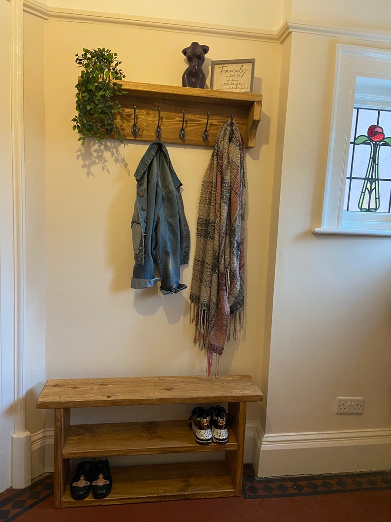 May include: A wooden coat rack with five hooks and a shelf. A denim jacket, a patterned scarf, and a decorative figurine are hanging on the hooks. The shelf has a sign that reads "Family is where life begins and love never ends". A wooden bench with two shelves is below the coat rack. The bench has two pairs of shoes on the bottom shelf.