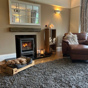 May include: A cozy living room with a wood-burning fireplace, a plush leather armchair with a furry throw pillow, and a dog bed with a dog sleeping in it. The room has a gray rug, a wooden shelf with logs, and a large mirror above the fireplace.