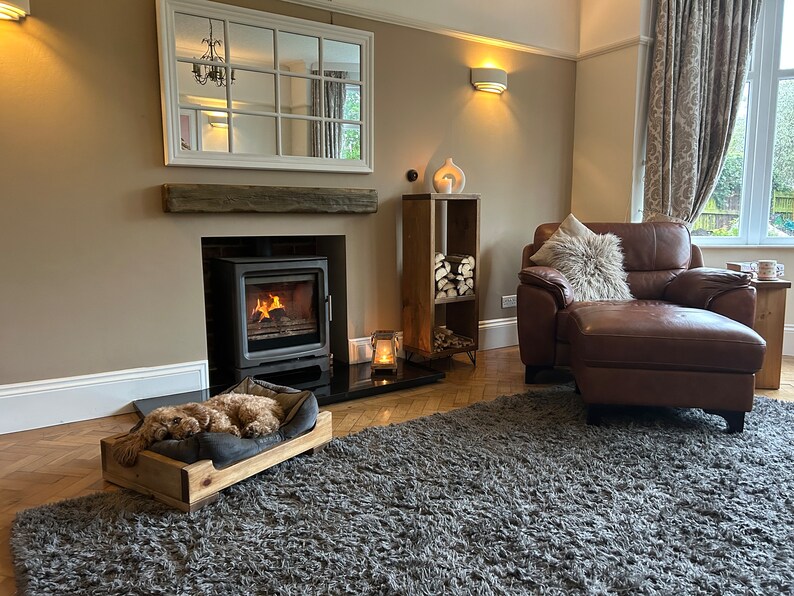 May include: A cozy living room with a wood-burning fireplace, a plush leather armchair with a furry throw pillow, and a dog bed with a dog sleeping in it. The room has a gray rug, a wooden shelf with logs, and a large mirror above the fireplace.