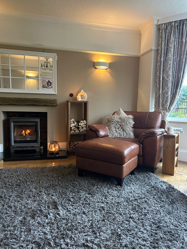May include: A brown leather armchair with a fluffy white throw pillow and a matching ottoman. The armchair is in front of a fireplace with a lit fire. A gray shag rug covers the floor.