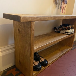 May include: A rustic wooden shoe rack with two shelves. The rack is made of natural wood with a light brown finish. There are shoes on the shelves, including a pair of black sandals and a pair of leopard print trainers.