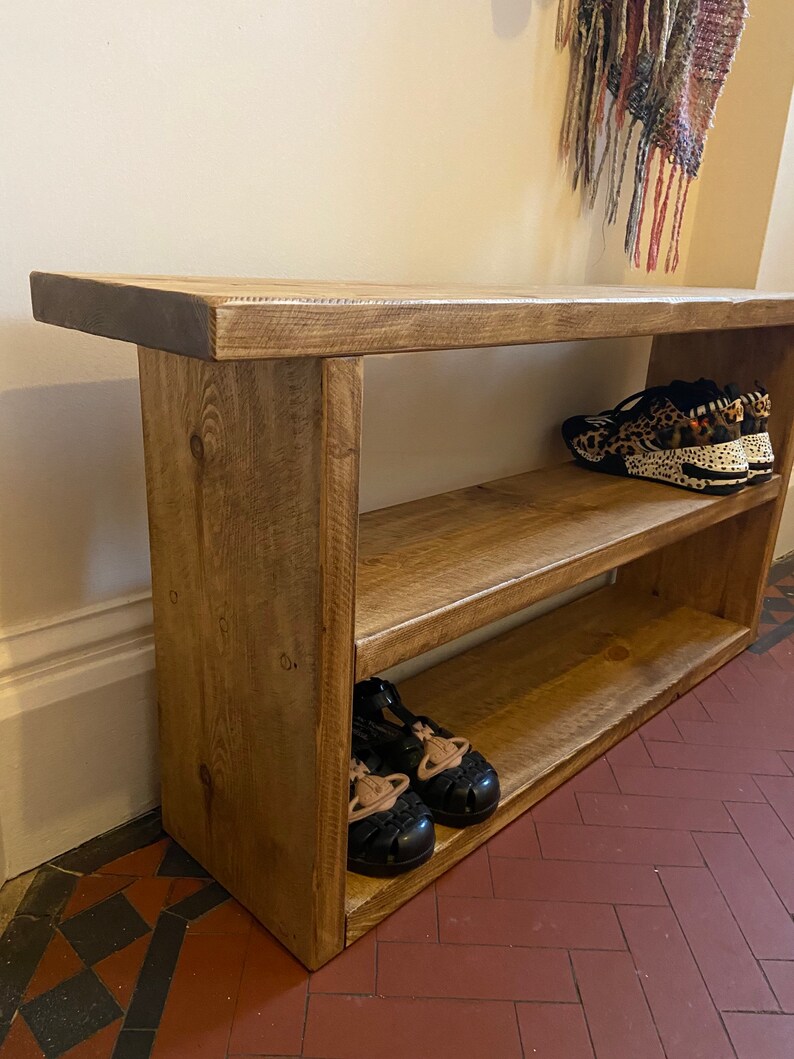 May include: A rustic wooden shoe rack with two shelves. The rack is made of natural wood with a light brown finish. There are shoes on the shelves, including a pair of black sandals and a pair of leopard print trainers.