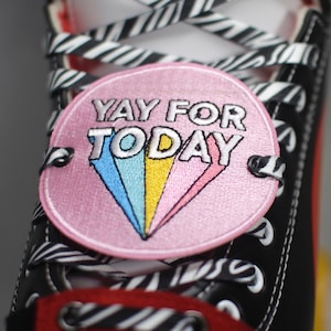 Könnte beinhalten: Nahaufnahme eines schwarzen und roten High-Top-Sneakers mit weißen Nähten und zebra-gestreiften Schnürsenkeln. Ein rosa Aufnäher mit dem Text "YAY FOR TODAY" und einem Regenbogen-Design ist am Schuh befestigt.