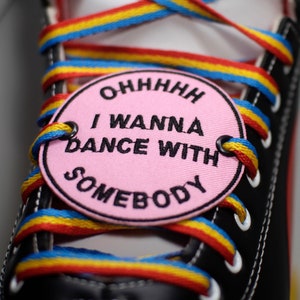 May include: Close-up of a black trainer with rainbow laces and a pink tag. The tag reads "OHHHH I WANNA DANCE WITH SOMEBODY" in black text. The laces are red, yellow, and blue.