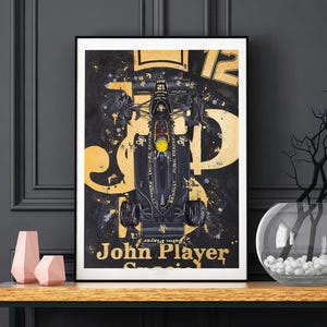 Op de afbeelding: Een ingelijste kunstprint met een bovenaanzicht van een zwarte en gouden Formule 1-raceauto. Het kunstwerk bevat de tekst "John Player Special" en het nummer "12" in een gouden lettertype tegen een zwarte achtergrond. De print staat op een houten tafel.