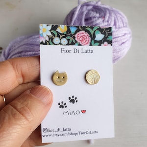 Peut inclure: Une paire de boucles d'oreilles en or en forme de chat et de pelote de laine sur une carte blanche avec le texte "Fior Di Latta" et "MIAO" avec un cœur.