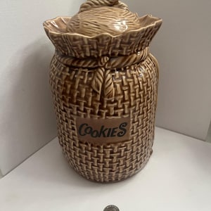 May include: A brown ceramic cookie jar with a woven basket design. The jar has a lid with a rope handle and the word "Cookies" on the front. The jar is a light brown color.