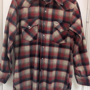 May include: A red, black, and tan plaid button-up shirt with two chest pockets and silver buttons. The shirt has a classic collar and long sleeves. The brand name is visible on the collar.