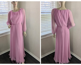 May include: A light pink vintage maxi dress with long, sheer sleeves and a pleated bodice and skirt. The dress has a round neckline and a fitted waist. The dress is displayed on a mannequin in front of a window.