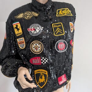 May include: Black sequined jacket adorned with patches featuring car brands like Ferrari, Jaguar, and Lamborghini. The jacket has a high collar and long sleeves. Patches include the text "Morgan", "Monitor", "Citroën", "Austin Healey", and "Uniroyal".