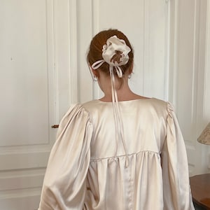 May include: A woman with long, red hair wearing a light beige satin blouse with puffed sleeves and a low neckline. She is wearing a beige satin hair scrunchie with a floral design in her hair.