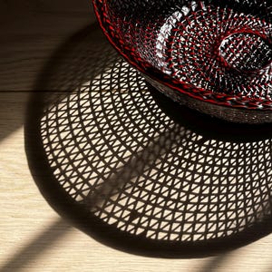 May include: A black wire basket with a red rim and a spiral design in the center. The basket casts a patterned shadow on a light-colored wooden surface, with sunlight creating highlights and shadows.