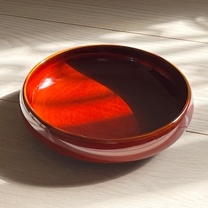 以下が含まれることがあります： 光沢のある赤茶色の仕上げが施された、丸い木製のボウル。ボウルの内側は光を反射し、その影は明るい色の木製の表面に落ちています。ボウルはシンプルでエレガントなデザインです。