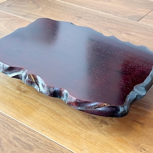 May include: A rectangular, dark burgundy wooden serving tray with a natural, wavy edge. The tray has a smooth, polished surface and sits on a light brown wooden surface. The edges are intricately carved.