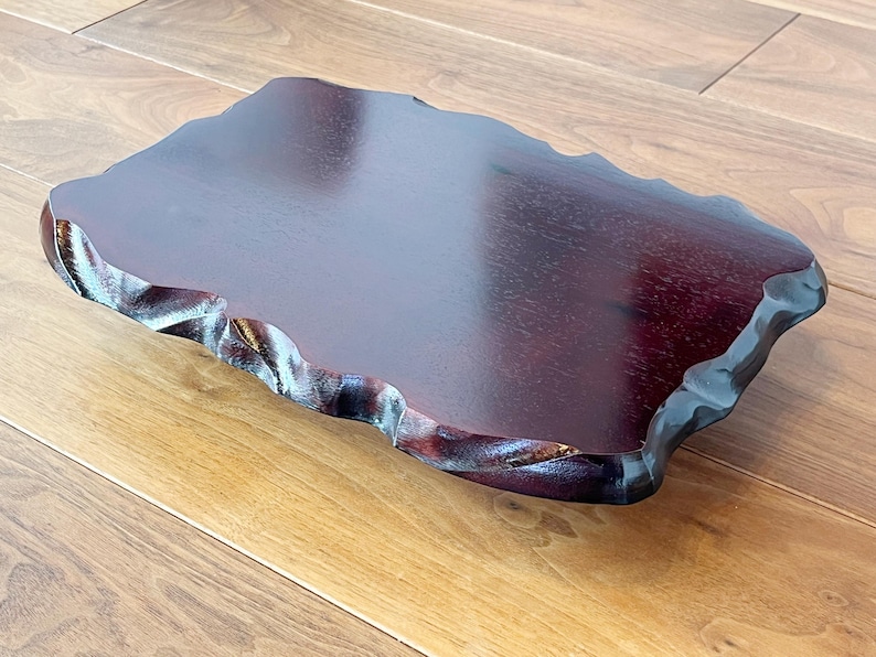 May include: A rectangular, dark burgundy wooden serving tray with a natural, wavy edge. The tray has a smooth, polished surface and sits on a light brown wooden surface. The edges are intricately carved.