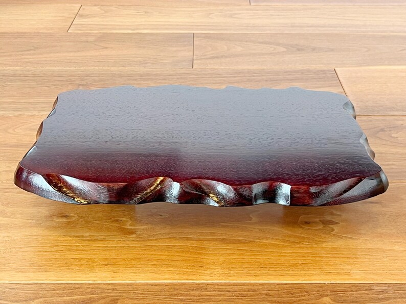 May include: A rectangular, dark red wooden tray with a natural, wavy edge. The tray has a glossy finish and appears to be made of a single piece of wood. The tray is approximately 30 cm long and 20 cm wide.