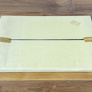 May include: A rectangular, light beige cardboard box, sealed with tan tape, rests on a wooden surface. The box is closed and appears to be empty. The wood grain is visible.