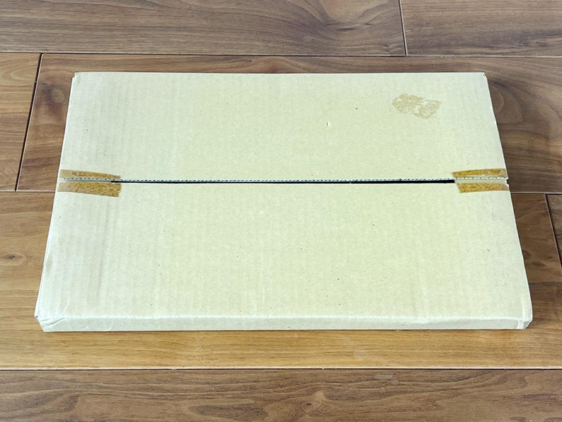 May include: A rectangular, light beige cardboard box, sealed with tan tape, rests on a wooden surface. The box is closed and appears to be empty. The wood grain is visible.