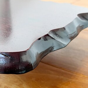 May include: Close-up of a wooden serving tray with a dark, glossy finish. The tray has a unique, wavy edge and a smooth, light-coloured surface. The wood grain is visible, adding texture to the design.