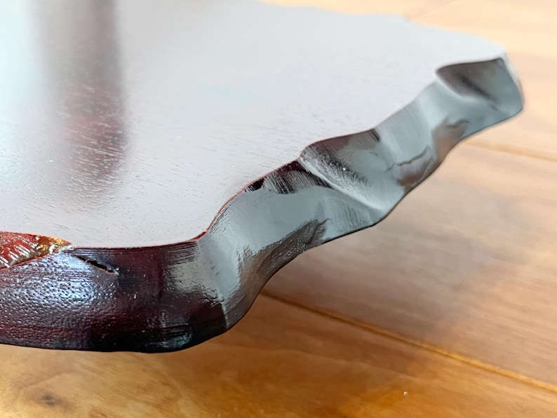 May include: Close-up of a wooden serving tray with a dark, glossy finish. The tray has a unique, wavy edge and a smooth, light-coloured surface. The wood grain is visible, adding texture to the design.