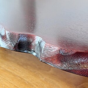 May include: Close-up of a wooden table edge with a dark reddish-brown finish. The edge has a decorative, wavy design with a lighter, textured accent. The table sits on a light brown surface.