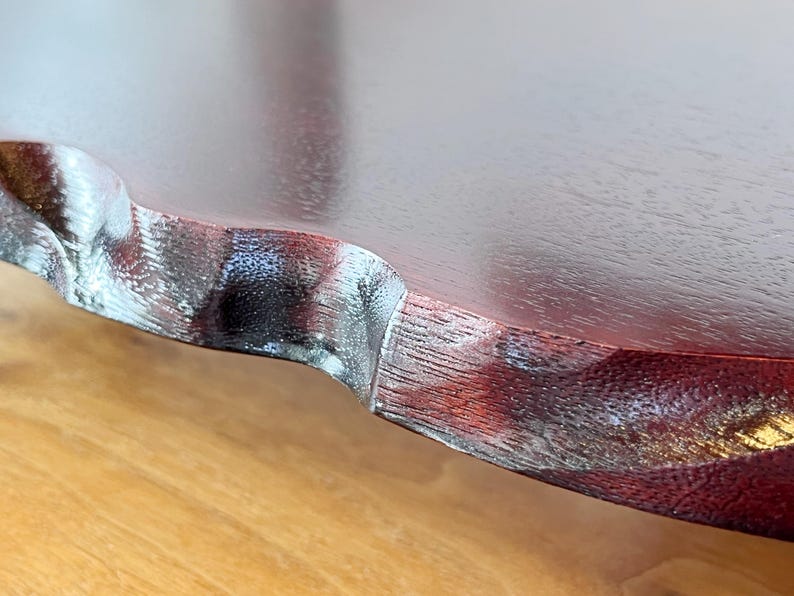 May include: Close-up of a wooden table edge with a dark reddish-brown finish. The edge has a decorative, wavy design with a lighter, textured accent. The table sits on a light brown surface.