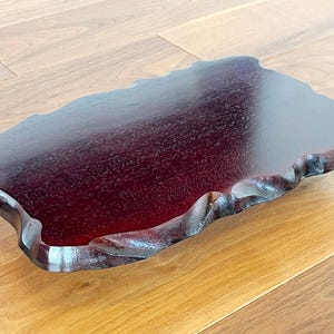 May include: A dark red wooden tray with a natural, irregular edge. The tray has a glossy finish and is displayed on a light brown wooden surface. The tray's unique shape and colour make it a decorative item.