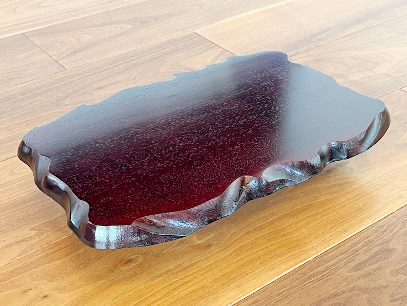 May include: A dark red wooden tray with a natural, irregular edge. The tray has a glossy finish and is displayed on a light brown wooden surface. The tray's unique shape and colour make it a decorative item.