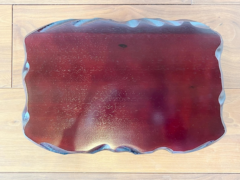May include: A rectangular, wooden serving tray with a deep burgundy finish. The edges have a wavy, scalloped design, and the tray sits on a light-coloured wooden surface. The tray's dimensions are not visible.